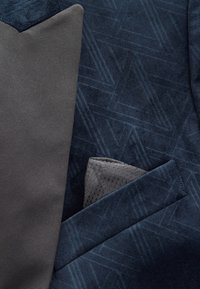 Veste en velours bleu foncé avec un motif géométrique subtil, une cravate gris côtelé et un mouchoir de poche gris texturé.