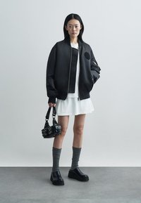 Črna bomberska jakna z okroglo našitkom, oblečena čez belo obleko. Dodatki vključujejo črno torbico, sivo nogavico in robustne črne čevlje.