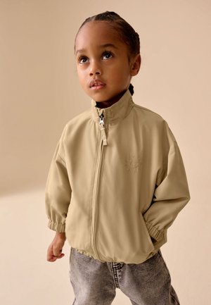 Lille barn med flettet hår iført beige lynlåstrøje og grå jeans, kigger opad mod en neutral baggrund.