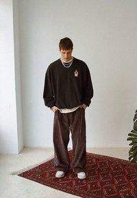Młody mężczyzna z krótkimi włosami, ubrany w warstwowy czarny sweatshirt, luźne ciemne spodnie i białe sneakersy, stoi na czerwonym, wzorzystym dywaniku w pomieszczeniu.