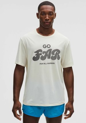Uomo che indossa una camicia bianca a maniche corte con il testo "GO FAR FOR ALL RUNNERS" e pantaloncini sportivi blu, in piedi su uno sfondo semplice.