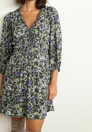 Femme portant une robe fleurie à col en V avec bordure à volants et taille froncée, associée à des bottes hautes taupe, debout devant un mur blanc uni.