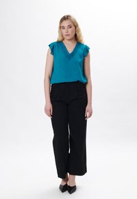 Blusa teal a volant con scollatura a v e motivo testurizzato, abbinata a pantaloni neri a vita alta con gamba larga e scarpe nere a punta.