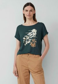 Donkergroen T-shirt met korte mouwen, versierd met bloemmotieven in goud en beige. Gecombineerd met lichtbeige broek.