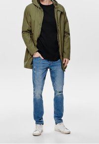 Man som bär en olivgrön huva jacka, svart skjorta, slitna blå jeans och vita sneakers, stående med en hand i fickan.