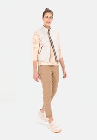 Leichte, gesteppte Weste in Off-White mit einem Reißverschluss vorne und braunen Streifen, getragen über einem gestreiften orange-weißen Oberteil und beigen Hosen, kombiniert mit hellen Sneakers.