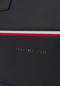 Navy-blue karhennettu nahkainen laukku, jonka alaosassa on vaakasuora punainen, valkoinen ja punainen raita sekä hopeinen "TOMMY HILFIGER" -logo.