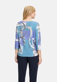 Blaue und weiße gemusterte Bluse mit ¾-Ärmeln, die Streifen und abstrakte Designs aufweist, aus einem leichten Stoff gefertigt.