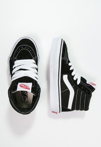 Czarne zamszowe sneakersy wysokie z białymi sznurówkami, białymi gumowymi podeszwami, bocznymi paskami i czerwoną metką z logo na języku. Teksturowana powierzchnia i szczegóły przeszycia.