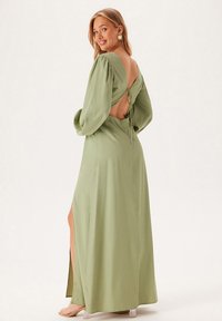 Dlouhé, olivově zelené šaty s nabíranými rukávy, otevřeným zadním dílem a zavazováním. Obsahují boční rozparek pro snadnější pohyb. Hladká textura.