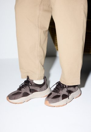 Piernas con pantalones beige que llevan zapatillas deportivas gruesas grises y negras con suelas blancas y calcetines blancos hasta el tobillo, de pie sobre una superficie blanca.
