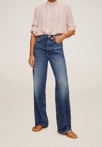 Helles rosa, gefaltetes Blusenshirt mit hohem Kragen, kombiniert mit hochtaillierten, weit geschnittenen blauen Jeans und braunen flachen Sandalen.