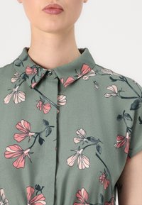 Blusa floreale verde con maniche corte, colletto abbottonato e motivi di fiori rosa su tessuto morbido.