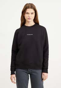 Fekete pamut pulóver hosszú ujjakkal, bordázott mandzsetta és kerek nyakú. A mellkason fehér "Calvin Klein Jeans" logó található.