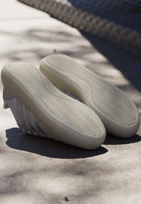 Paire de baskets blanches avec des semelles en caoutchouc épaisses et texturées reposant sur une surface en béton ensoleillée près d'une clôture grillagée.