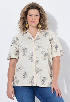 Kvinde med krøllet hår iført øreringe formet som gyldne blade, en cremet kortærmet skjorte med blomsterprint og blå jeans, stående op ad en hvid væg.