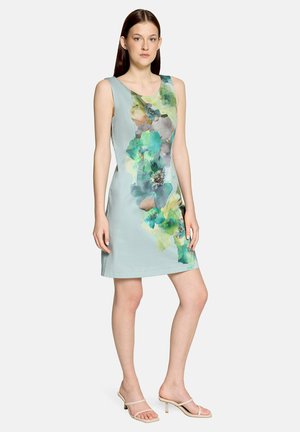 Femme debout portant une robe sans manches bleu clair avec de grandes impressions florales vertes et bleues et des sandales beiges à talons ouvert.