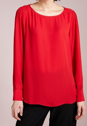 Frau trägt eine knallrote Bluse mit langen Ärmeln und plissierten Schulterdetails sowie schwarze Hose, steht vor einem schlichten Hintergrund.
