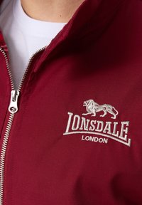 Bordo zip-up jakna s izvezenim logotipom "Lonsdale London" i dizajnom lava. Ima glatku teksturu i srebrne metalne dijelove na patentnom zatvaraču.