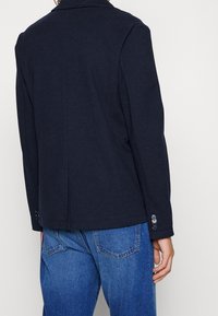 Marinblå bomullsblazer med skräddarsydd design, slagkrage och knäppte manschetter. Synlig sömnadsdetalj och mjuk textur.