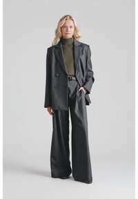 Kobieta z długimi blond włosami, ubraną w ciemnoszary oversize'owy żakiet i spodnie o szerokich nogawkach oraz oliwkowy sweter z golfem, stojąca z jedną ręką w kieszeni.