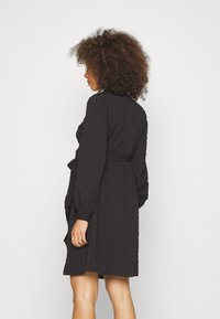 Vestido preto com um tecido texturizado e enrugado, mangas longas, cintura com laço e um corte relaxado, caindo acima dos joelhos. Design simples.