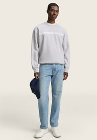 Pilkas džemperis su baltu juostele, šviesiai mėlynos džinsai, ir balti sportbačiai. Laikantis tamsiai mėlynos beisbolo kepurės, stovi priešais vientisą fono.