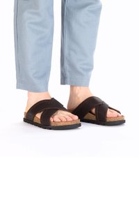 Bruine leren sandalen met kruislings banden, kurken voetbed, structuur van zwart rubberen zool en open teen ontwerp. Gedragen met lichtblauwe broek.