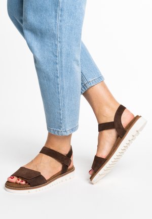 Brown suede sandals with wide straps, white rubber sole, textured footbed, and open-toe design. Paired with light blue cropped jeans.