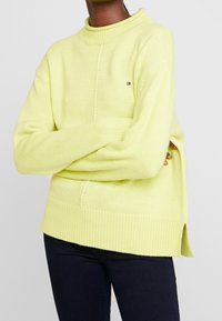 Sweter w kolorze limonkowym, wykonany na drutach, z wysokim dekoltem, szerokimi rękawami, bocznymi rozcięciami i małym logo na piersi. Teksturalna i przytulna tkanina.