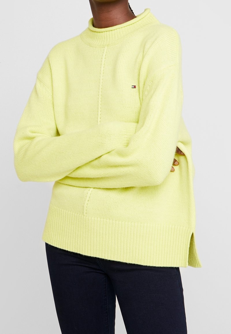 Sweter w kolorze limonkowym, wykonany na drutach, z wysokim dekoltem, szerokimi rękawami, bocznymi rozcięciami i małym logo na piersi. Teksturalna i przytulna tkanina.