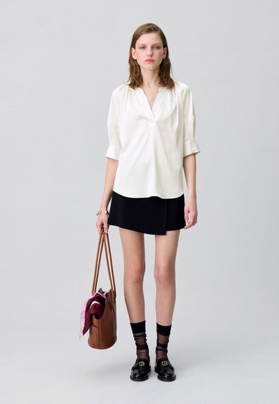 Camisa blanca de manga corta con escote en V, combinada con una falda negra. El modelo sostiene un bolso marrón y lleva mocasines negros con medias finas.