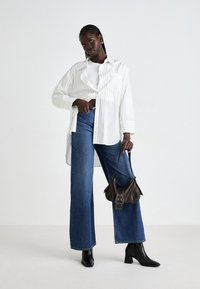 Donna con capelli intrecciati che indossa jeans larghi blu, top corto bianco, camicia bianca a righe larghe, stivali neri e tiene in mano una piccola borsa marrone.