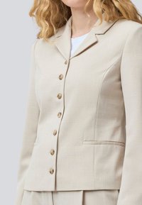 Beige skräddarsydd blazer med notch-krage, enkelknäppt design, sex knappar och två framfickor. Slät tygkvalitet.