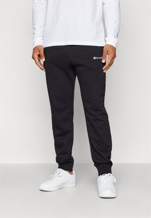 Pantalones deportivos negros con puños elásticos y un pequeño logo rojo y blanco en el lado izquierdo, combinados con zapatillas deportivas blancas.