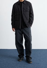 Zwarte lichte jas met vier voorzakken, gecombineerd met donkere cargobroek en zwarte sneakers. De stof lijkt glad met een relaxte pasvorm.