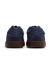 Granatowe zamszowe sneakersy z teksturowanym, wyściełanym granatowym piętą i zielonymi akcentami. Brązowa gumowa podeszwa ma przeszycie wokół krawędzi.