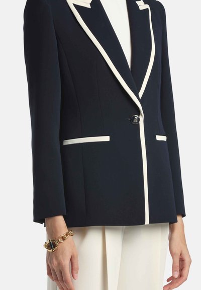 Námornícky blejzer s bielym lemovaním, zapínaný na jedno gombík a s dvoma prednými vreckami. Má hladkú textúru a priliehavý strih, vyrobený zo tkaniny.