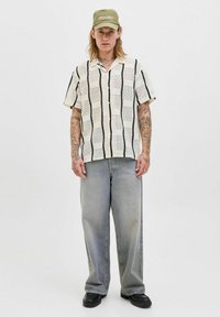 Chemise à manches courtes, de couleur crème, avec un motif à carreaux noir, associée à un jean en denim gris décontracté et une casquette verte. Tissu texturé avec des détails raffinés.