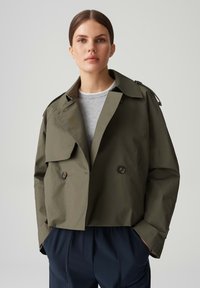 Olijfgroene cropped jas van lichtgewicht materiaal, met een brede kraag, twee voorkanten zakken en donkere knopen, gedragen over een grijs shirt.