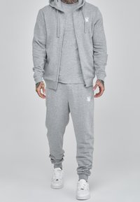 Sudadera gris y pantalones de chándal a juego, hechos de una tela suave, con cierre frontal de cremallera y detalle de logo, combinados con zapatillas blancas.