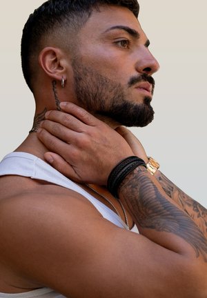 Black leather bracelets and a gold watch adorn a muscular arm. The person wears a white tank top and has a short, styled beard with tattoos.