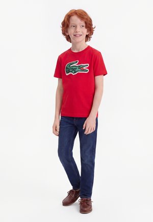 Garçon roux souriant portant un t-shirt rouge avec un logo de crocodile vert, un jean bleu et des chaussures marron à lacets, debout sur un fond blanc.