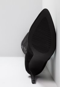 Bottine noire en suède à bout pointu avec un talon haut et fin, semelle tournée vers l'avant sur un fond blanc et gris.