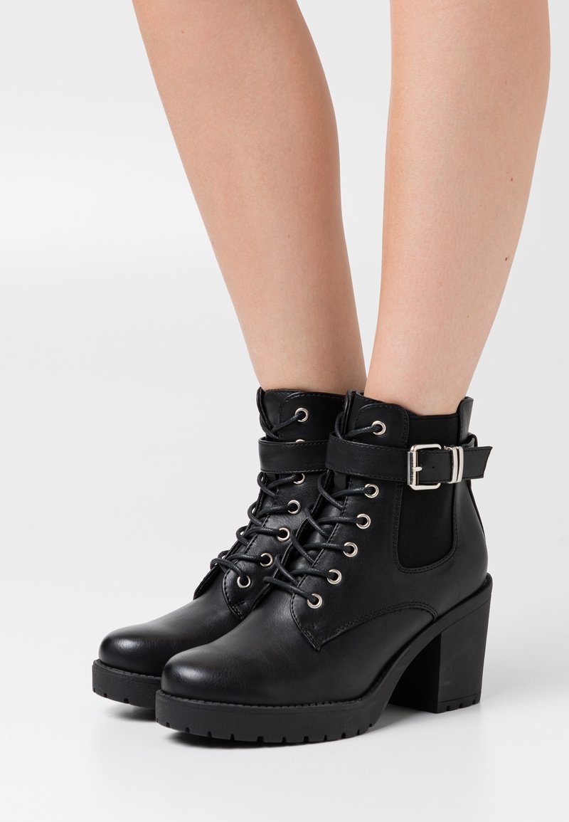 Bottines noires à lacets avec talons épais et brides à boucle latérale portées sur des jambes nues sur un fond uni.
