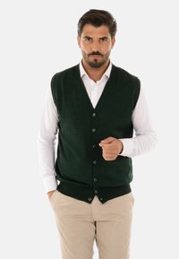 Gilet lavorato a maglia verde scuro con scollo a V, sei bottoni sul davanti, indossato sopra una camicia bianca, abbinato a pantaloni beige.
