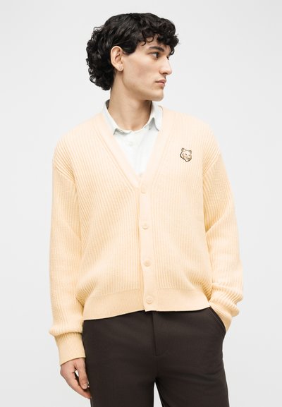 Νεαρός με σγουρά μαλλιά που φοράει εκρού ζακέτα με κουμπιά και ένα σήμα λυκόφωτος, πάνω από λευκό πουκάμισο και σκούρο παντελόνι, κοιτάζει προς τα δεξιά.