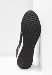 Semelle de chaussure de sport noire en caoutchouc texturé, avec des rainures horizontales et verticales prononcées, complétée par un détail de bord de semelle extérieure blanc.