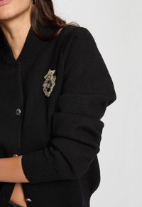 Cárdigan negro de punto con un collar de canalé, botones plateados y un broche decorativo con un detallado intrincado.