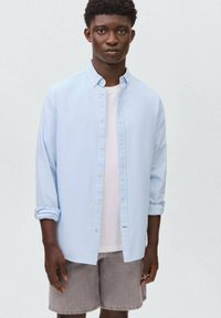 Un giovane uomo si erge con sicurezza, indossando una camicia a righe blu chiaro sopra una maglietta bianca e pantaloni corti grigi, su uno sfondo neutro.